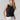 A person with long blonde hair is wearing the Twisted Tank Black by Sanctuary Clothing and white pants. They have their hands in their pockets and are looking directly at the camera with a neutral expression, set against a plain, light-colored background.