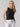 A person with long blonde hair is wearing the Twisted Tank Black by Sanctuary Clothing and white pants. They have their hands in their pockets and are looking directly at the camera with a neutral expression, set against a plain, light-colored background.