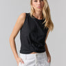 A person with long blonde hair is wearing the Twisted Tank Black by Sanctuary Clothing and white pants. They have their hands in their pockets and are looking directly at the camera with a neutral expression, set against a plain, light-colored background.