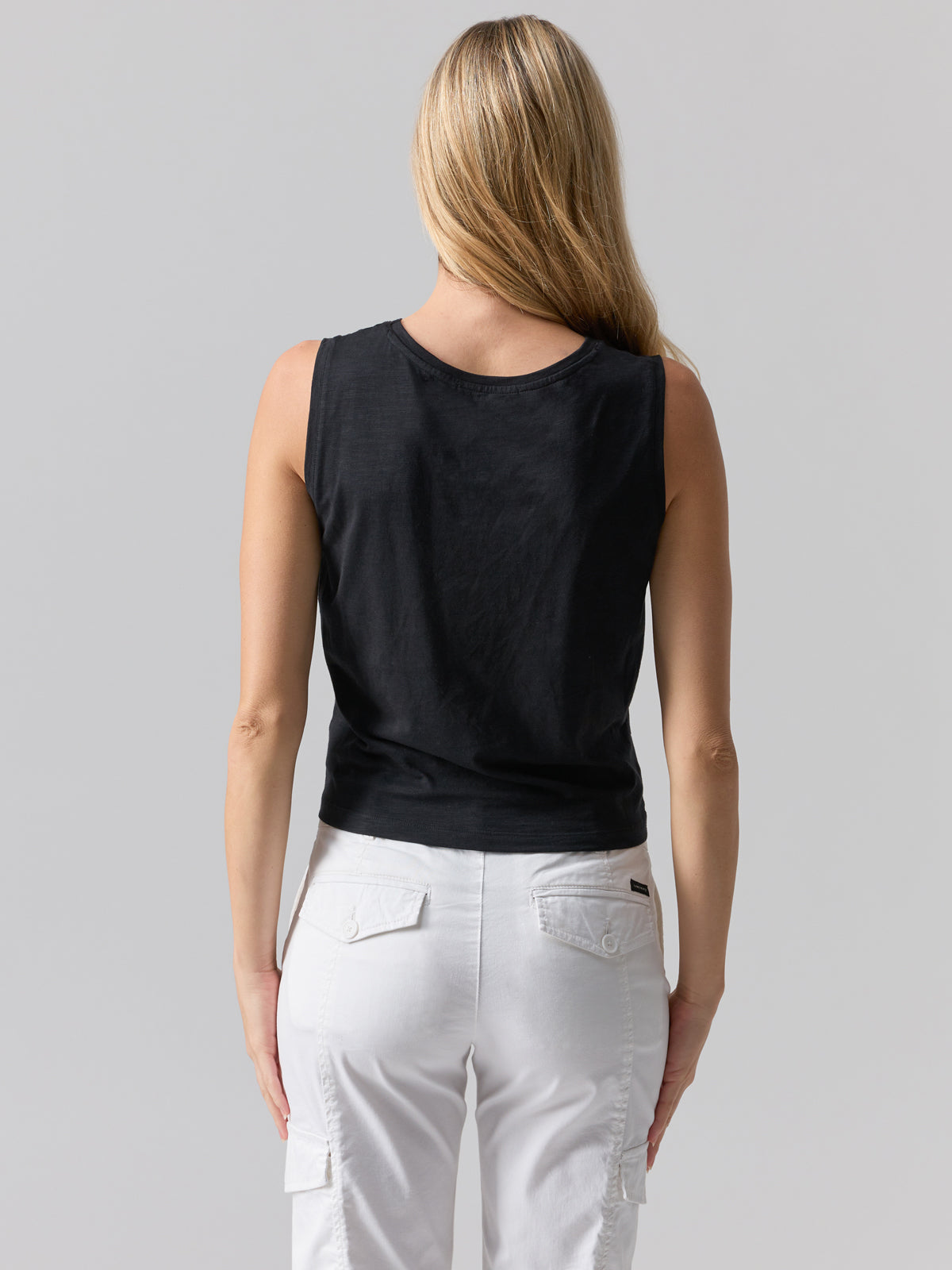 A person with long blonde hair is seen from the back, wearing a black Twisted Tank by Sanctuary Clothing and white pants with side pockets. The background is plain and neutral.