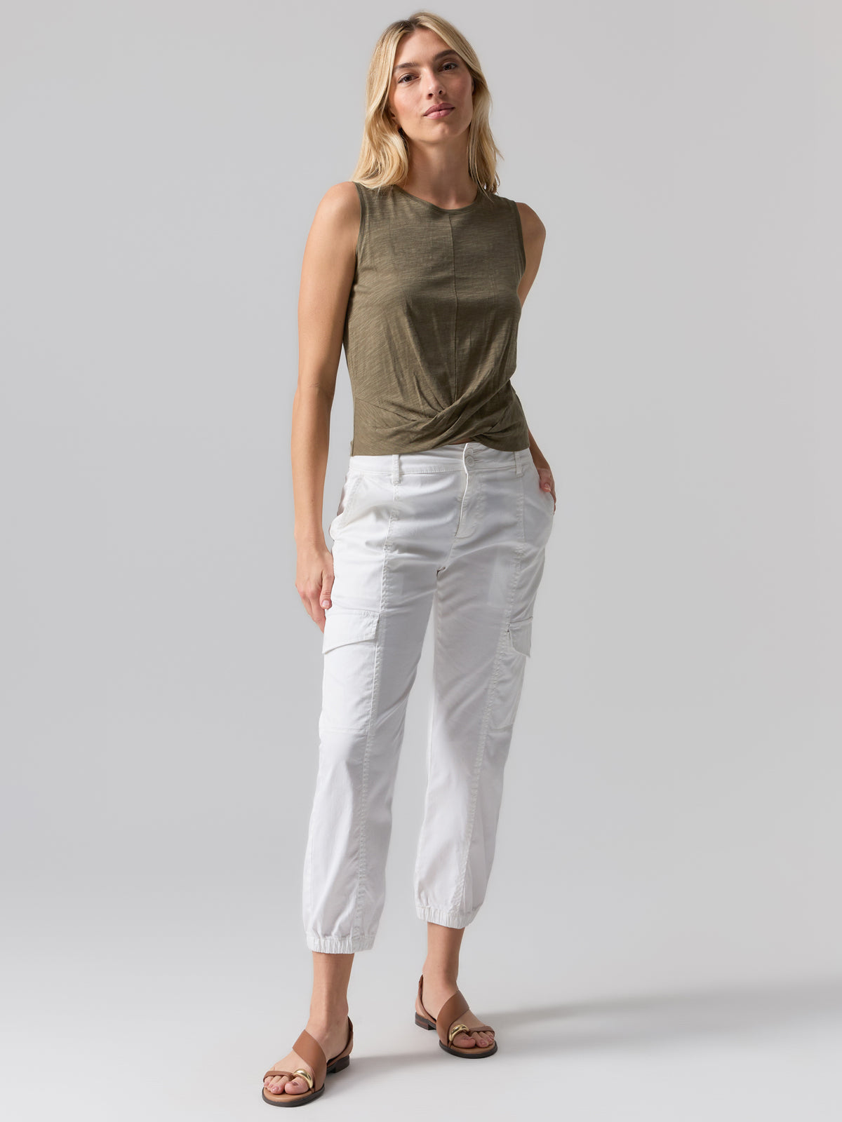 A person with shoulder-length blonde hair is wearing the Twisted Tank Burnt Olive by Sanctuary Clothing and white cropped cargo pants with an elastic hem. They are standing against a plain gray background with hands in the pockets, and wearing brown sandals.