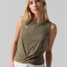 A woman with long blonde hair is wearing a Sanctuary Clothing Twisted Tank in Burnt Olive, paired with white pants. She is standing against a plain light grey background, looking at the camera with a neutral expression and her right hand in her pocket.