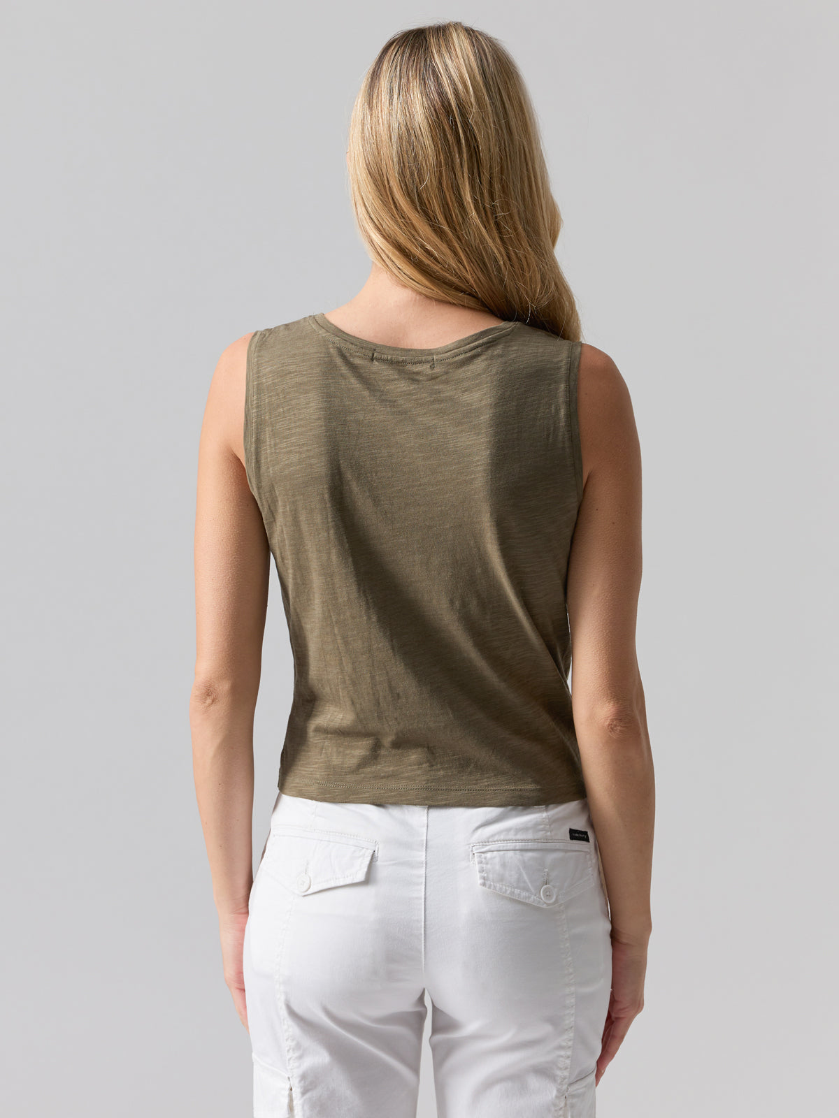 A person with long blonde hair is standing with their back to the camera, wearing the Sanctuary Clothing Twisted Tank in Burnt Olive and white pants that feature pockets and seams. The background is plain and light gray.