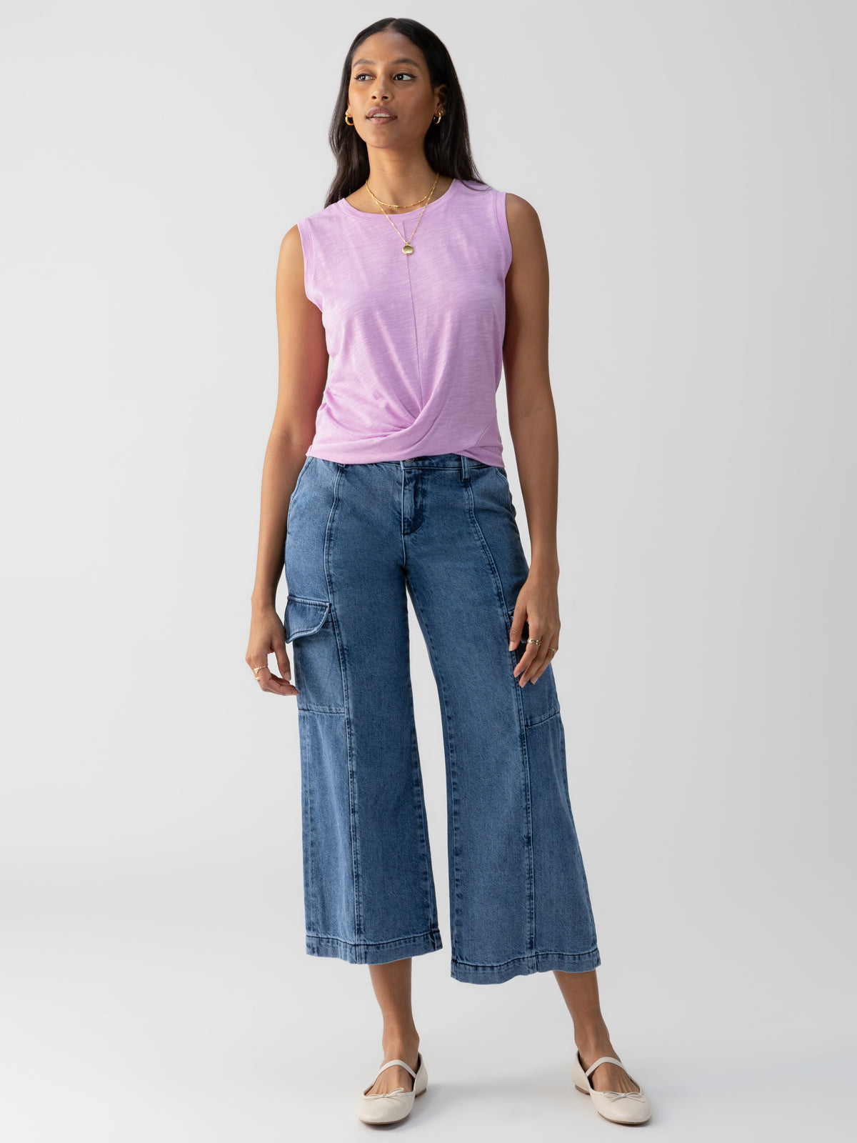 A woman stands against a plain background wearing the Twisted Tank Iris by Sanctuary Clothing, a sleeveless light purple top with a twist detail at the front. She pairs it with high-waisted, wide-leg denim pants featuring patch pockets, and completes the look with beige flats and a simple pendant necklace.