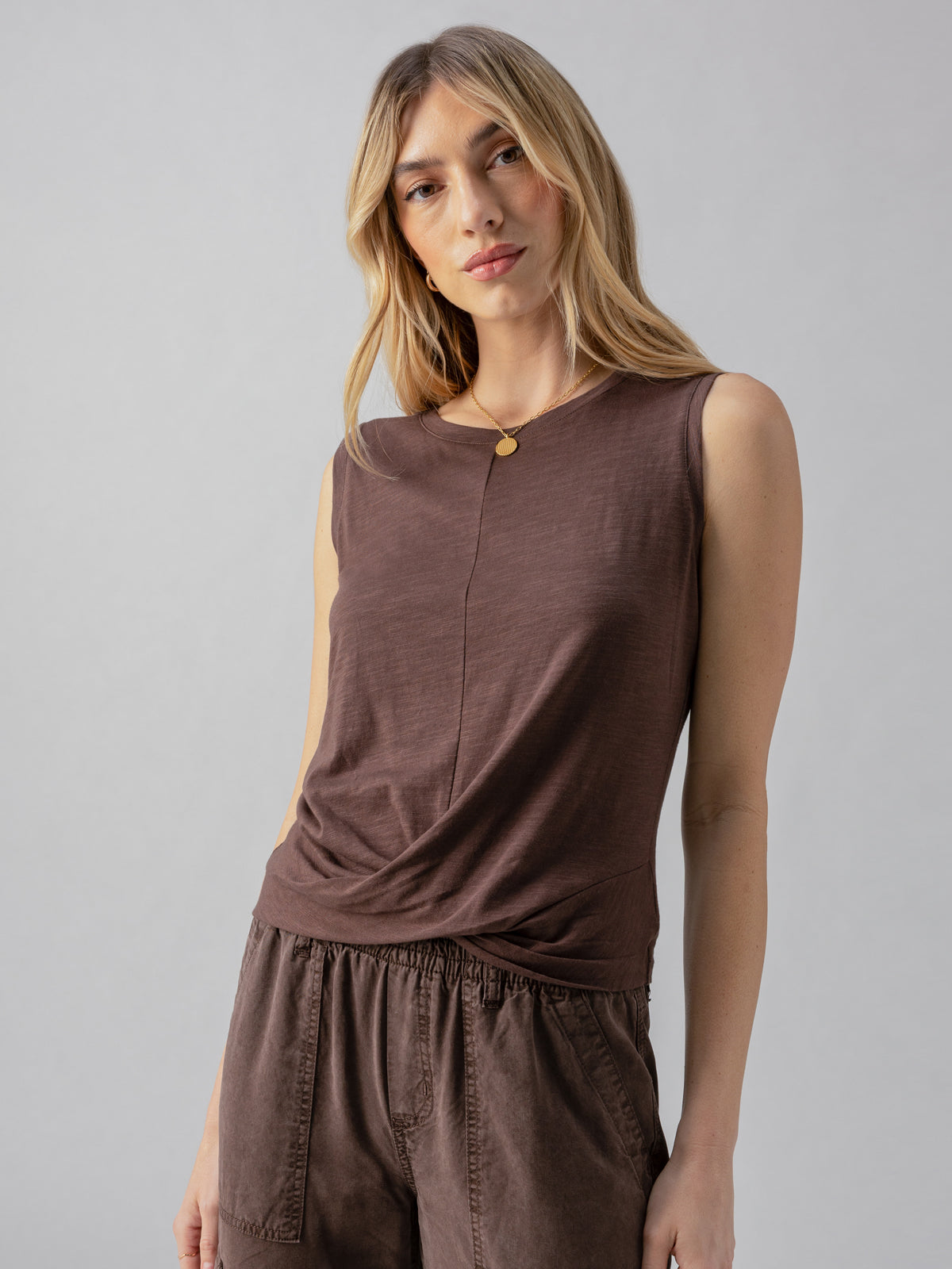 A person with long blonde hair wearing a Twisted Tank Mud Bath sleeveless top and matching brown pants from Sanctuary Clothing stands against a plain gray background. They have a relaxed pose with a slight smile, and there is a gold necklace visible around their neck.