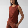 A woman with long dark hair stands against a plain background. She wears the Twisted Tank Rich Clay sleeveless top from Sanctuary Clothing, paired with matching rust-colored pants that have pockets. She has a calm expression, one hand in her pocket while the other rests by her side.