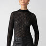 A person with their hair pulled back is wearing the "Long Sleeve Lace Mock Black" top from Sanctuary Clothing over black high-waisted pants. They are also adorned with small hoop earrings and posing against a plain white background.