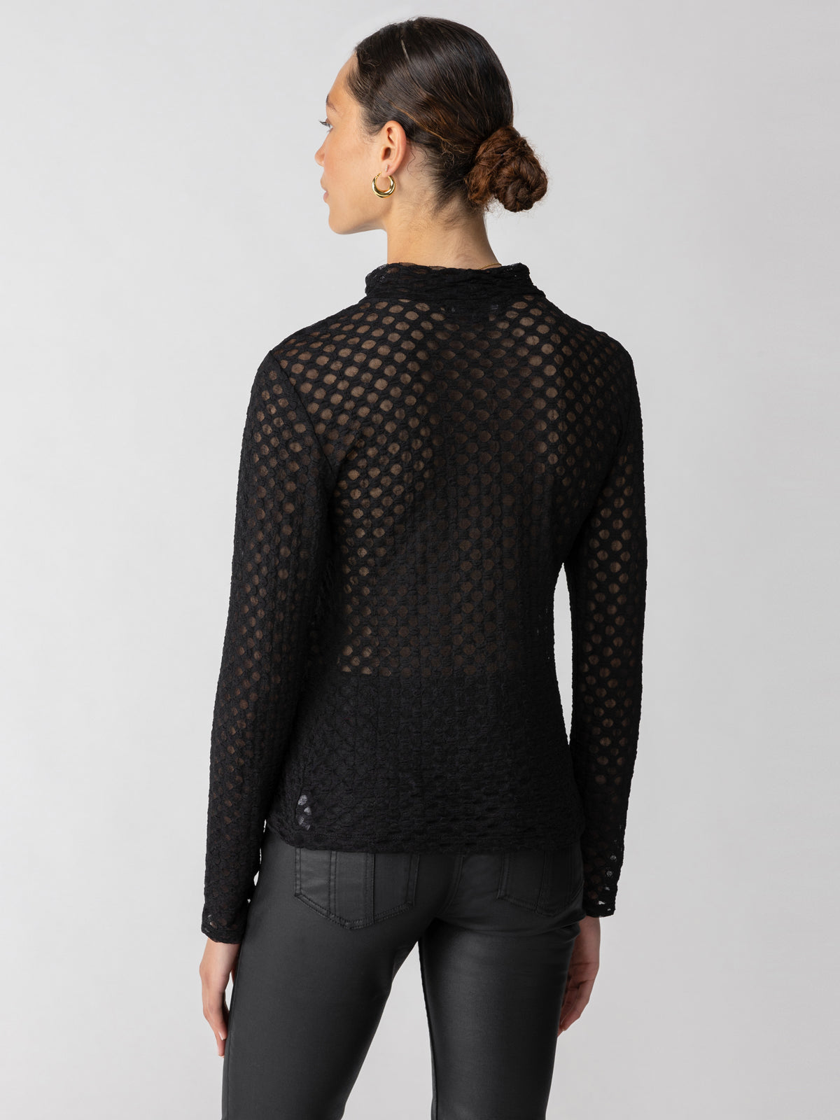 A person with dark hair tied back is seen from behind, wearing the Long Sleeve Lace Mock Black top by Sanctuary Clothing. The semi-transparent top features a hexagonal mesh pattern and long sleeves. The person is also wearing dark, fitted pants, set against a plain, light-colored background.