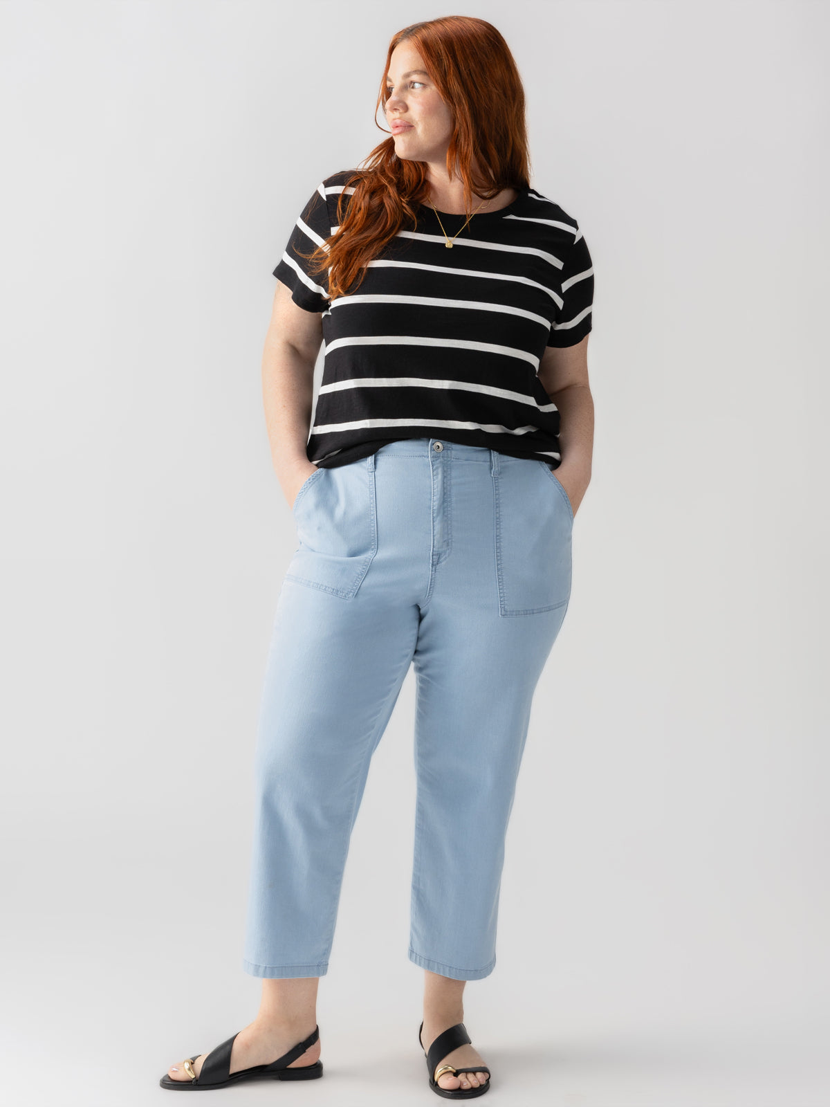 A woman with long red hair stands confidently against a gray background. She is wearing The Perfect Tee Black Stripe from the Inclusive Collection by Sanctuary Clothing, light blue high-waisted pants, and black sandals. Her hands are in her pockets, and she is looking off to the side with a relaxed expression.