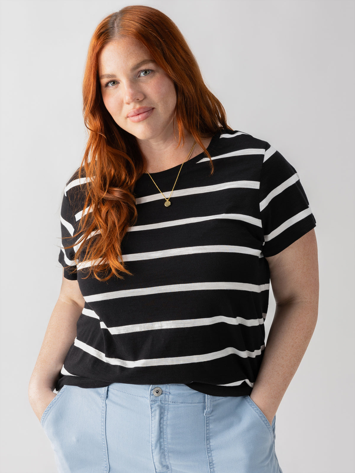 A woman with long, red hair is standing against a plain background, dressed in the Sanctuary Clothing's The Perfect Tee Black Stripe from the Inclusive Collection and light blue pants. She is smiling slightly with her hands in her pockets and accessorized with a gold necklace.