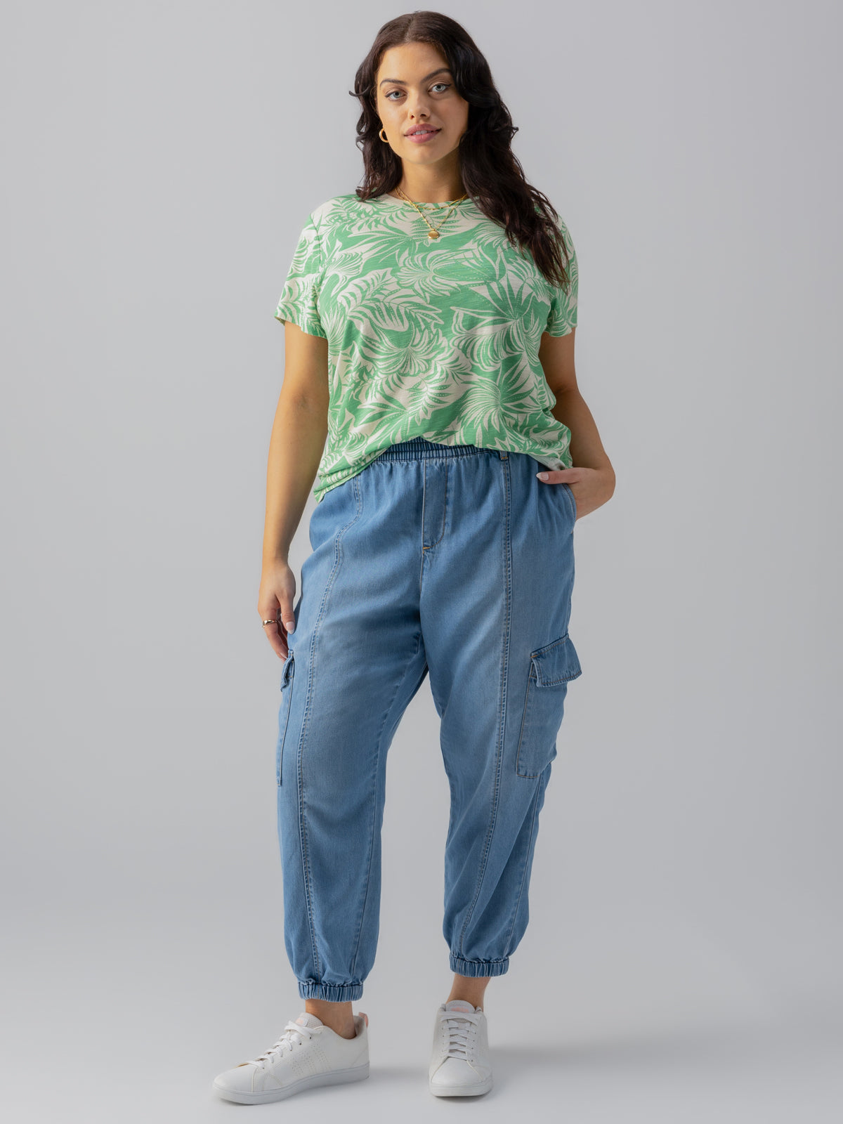 A person with long dark hair is standing against a plain background, wearing The Perfect Tee Cool Palm from Sanctuary Clothing's Inclusive Collection. They have paired it with light blue cargo pants featuring elastic cuffs and white sneakers. Sporting a relaxed expression, they have one hand in their pocket.