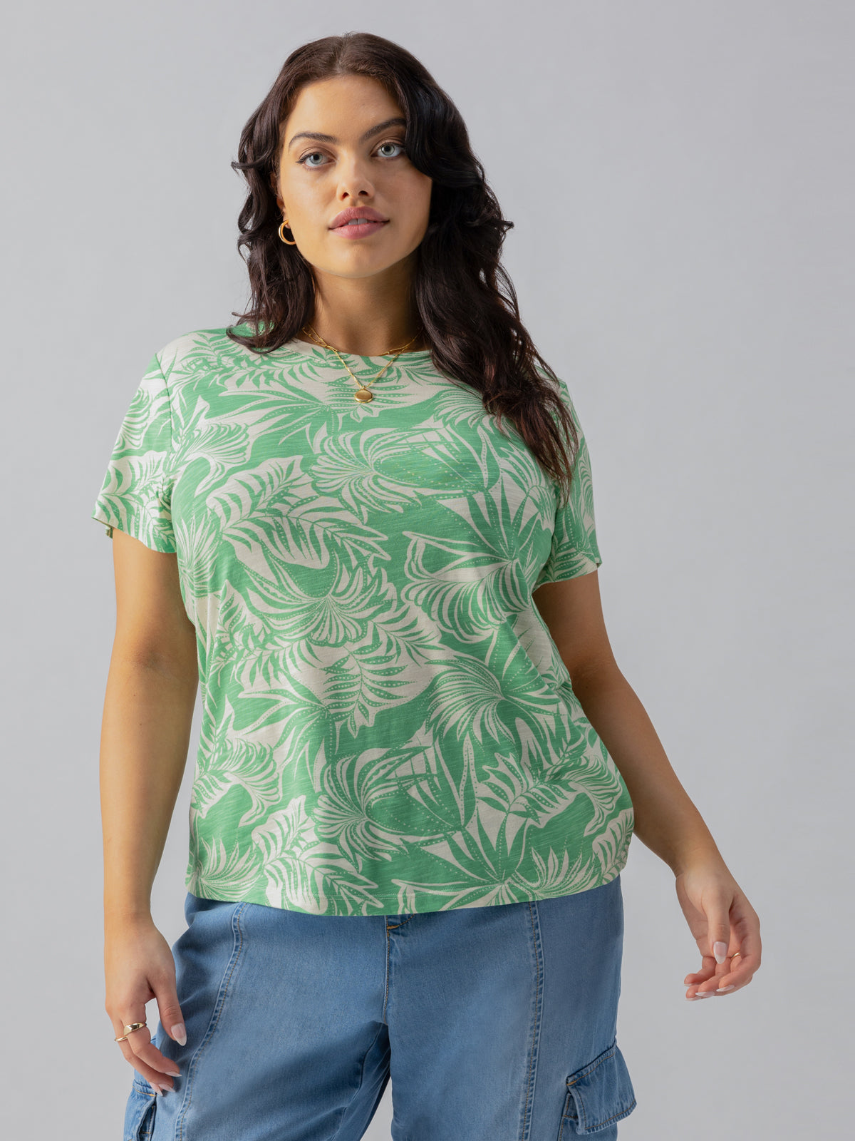 A woman with long dark hair wears The Perfect Tee Cool Palm Inclusive Collection from Sanctuary Clothing and light blue cargo pants. She stands against a plain gray background, gazing forward with a neutral expression. She is accessorized with gold hoop earrings and a gold necklace.