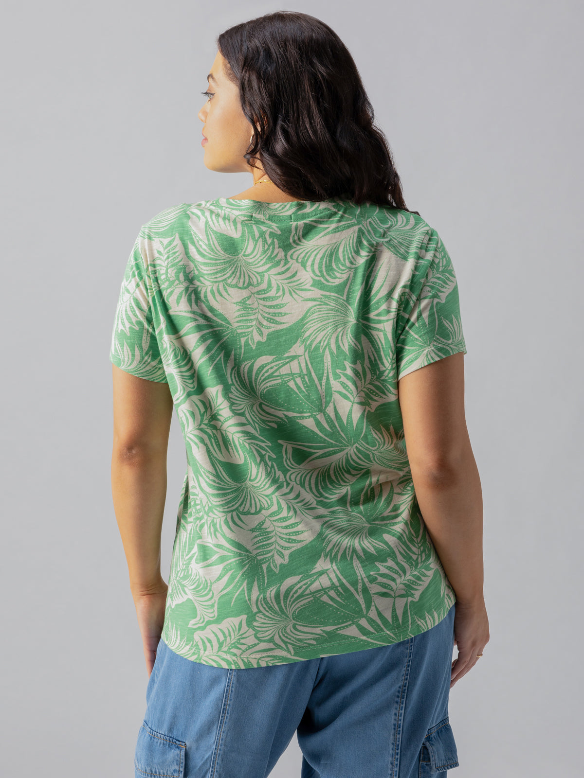 A person with long dark hair is seen from the back, wearing "The Perfect Tee Cool Palm Inclusive Collection" by Sanctuary Clothing, paired with blue cargo pants. The background is plain and light gray.