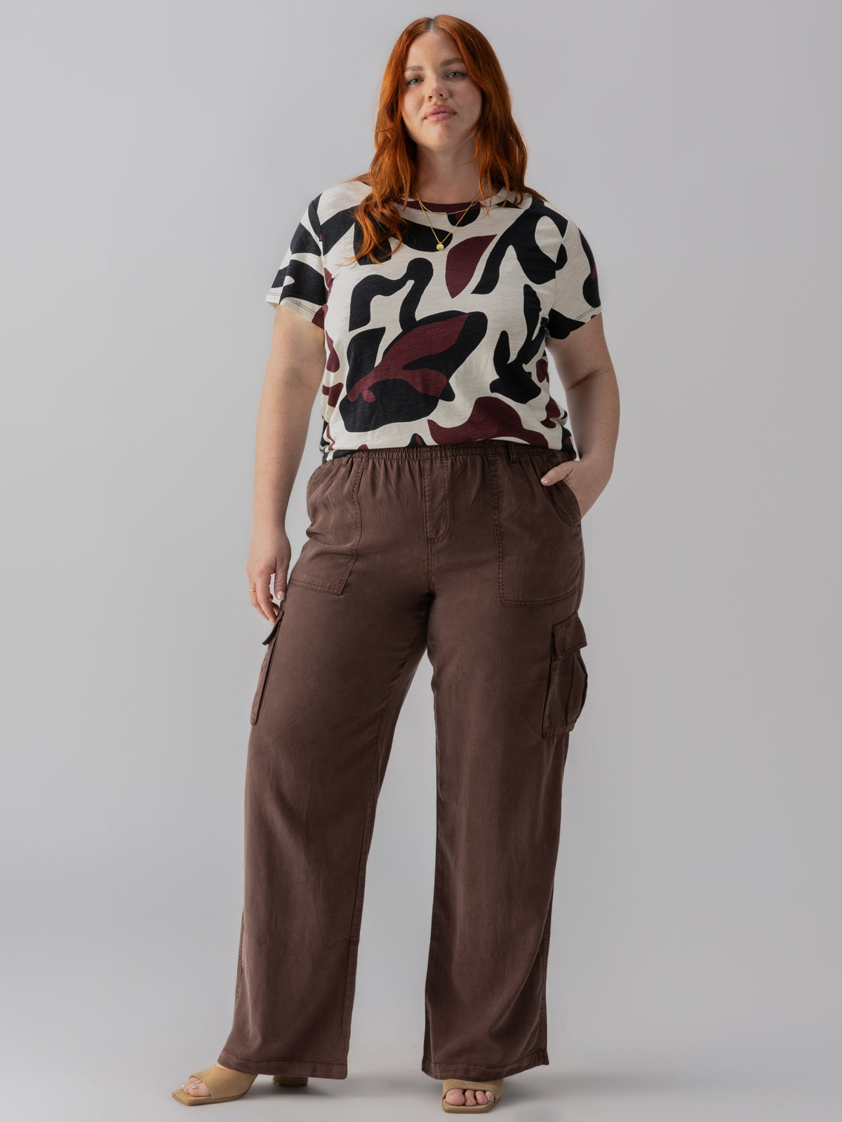 A person with long red hair stands against a gray background, wearing the Relaxed Reissue Mud Bath Inclusive Collection from Sanctuary Clothing. They don a short-sleeved, abstract-patterned shirt in black, white, and maroon, paired with loose brown pants featuring cargo pockets. With a relaxed and confident pose, they have one hand casually placed in their pocket.