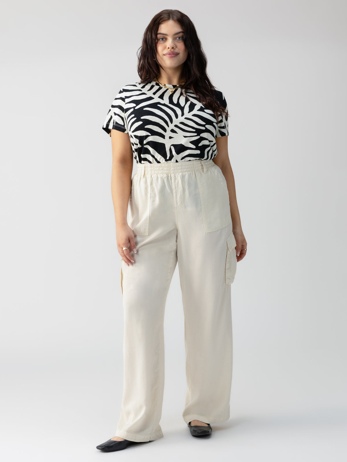 A woman with long brown hair stands against a white background. She is wearing The Perfect Tee Night Palm from the Inclusive Collection by Sanctuary Clothing and high-waisted, loose-fitting beige pants with pockets. She has black flats on her feet and is looking directly at the camera with a neutral expression.