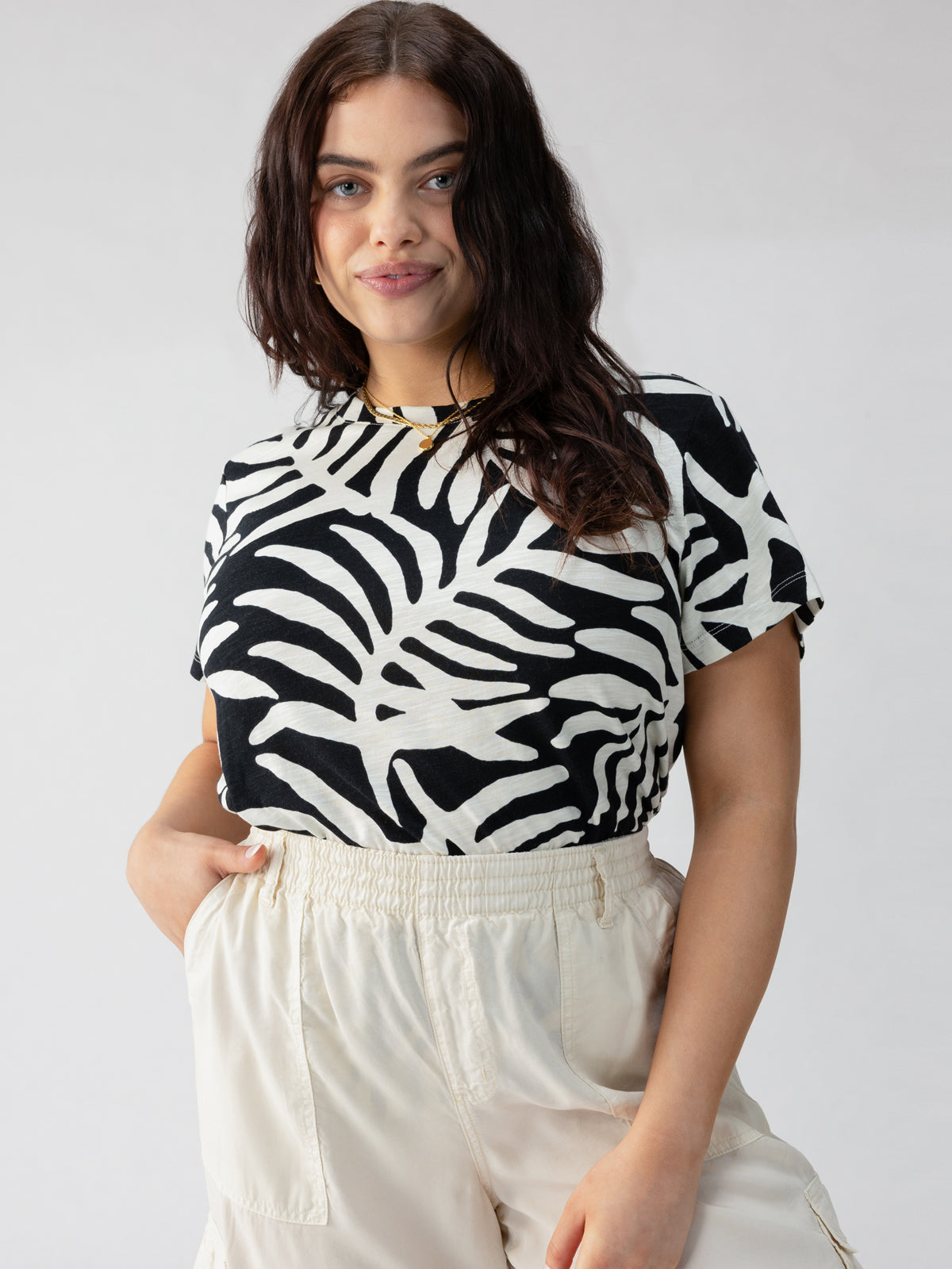 A person with long, wavy dark hair softly smiles while posing in Sanctuary Clothing’s The Perfect Tee Night Palm Inclusive Collection t-shirt and light-colored high-waisted pants. The background is plain and neutral, drawing focus to the person and their outfit.