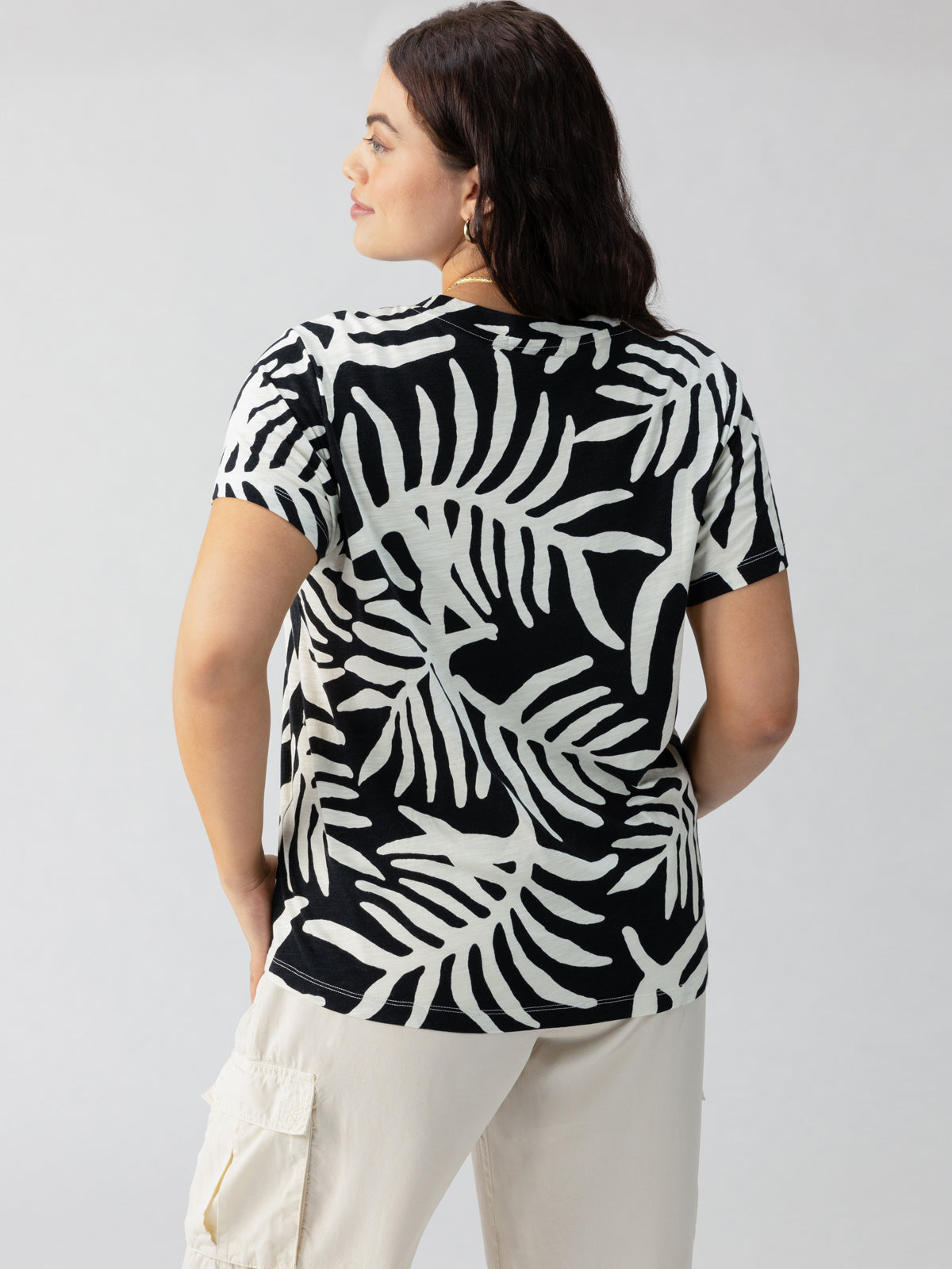 A person with long dark hair, seen from the back, is wearing Sanctuary Clothing's The Perfect Tee Night Palm Inclusive Collection, a black and white short-sleeved shirt featuring a large leaf pattern. The person is also dressed in light-colored cargo pants. The background is plain gray.