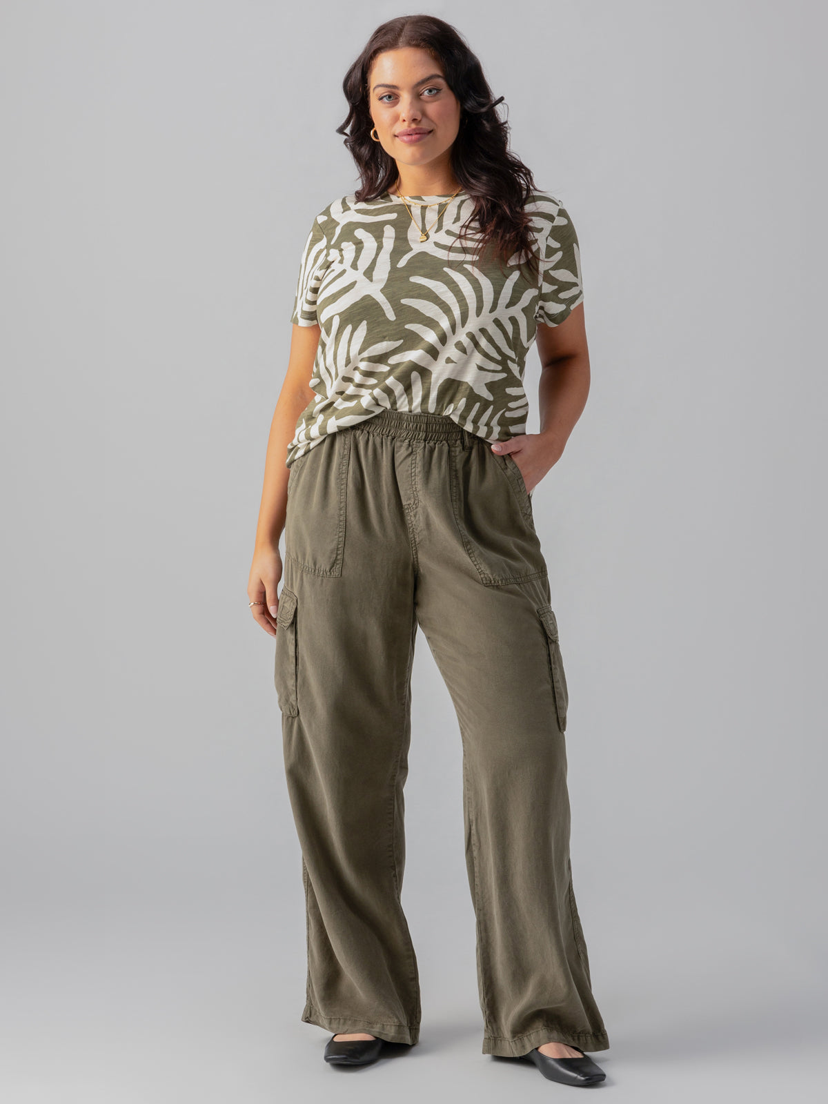 A woman stands against a plain background, wearing The Perfect Tee Olive Night Palm from the Sanctuary Clothing Inclusive Collection and olive green cargo pants. She has one hand in her pocket and is smiling slightly, with long, wavy hair. She is also wearing black shoes.