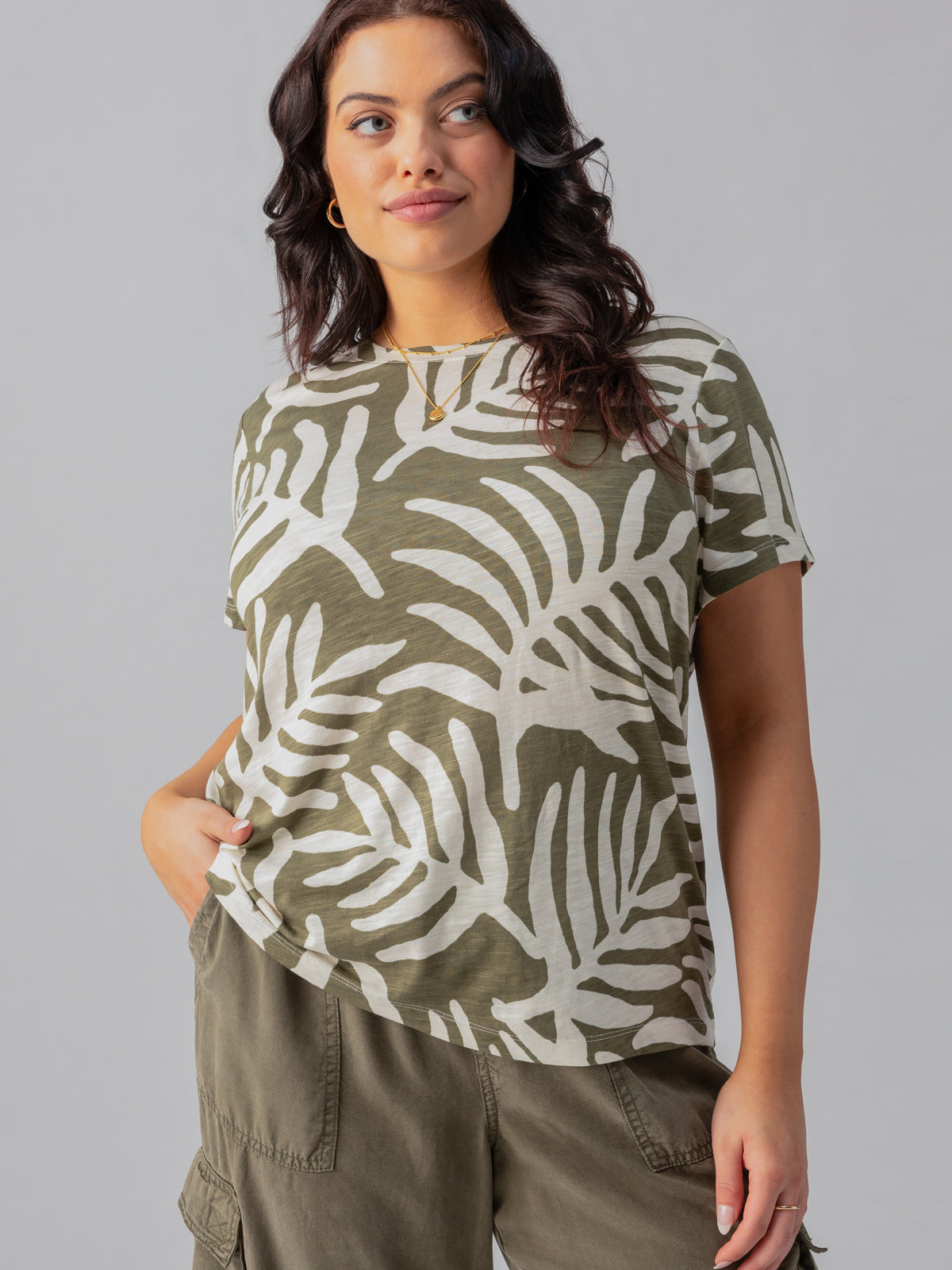 A woman with wavy dark hair is wearing "The Perfect Tee Olive Night Palm" from the Inclusive Collection by Sanctuary Clothing, along with olive cargo pants. She is standing with one hand in her pocket and the other relaxed by her side, looking slightly to her left with a soft smile. She is accessorized with a necklace.