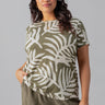 A woman with wavy dark hair is wearing "The Perfect Tee Olive Night Palm" from the Inclusive Collection by Sanctuary Clothing, along with olive cargo pants. She is standing with one hand in her pocket and the other relaxed by her side, looking slightly to her left with a soft smile. She is accessorized with a necklace.