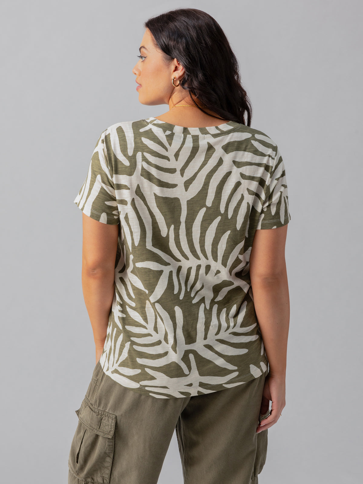 A woman with long, dark hair is standing with her back to the camera. She is wearing The Perfect Tee Olive Night Palm Inclusive Collection by Sanctuary Clothing, paired with olive green cargo pants. The background is a plain, light gray.