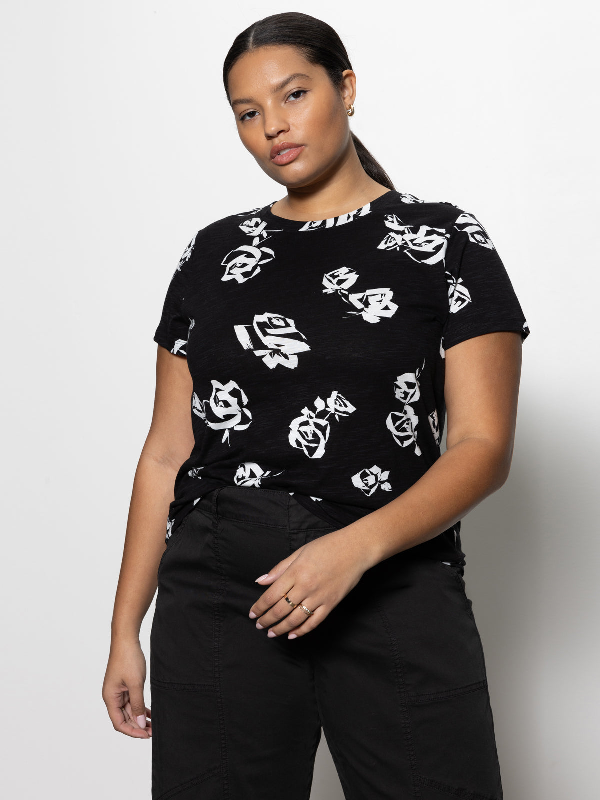 A woman wearing "The Perfect Tee" from the Rose Pop Inclusive Collection by Sanctuary Clothing stands against a plain background. She has her hair pulled back and is wearing black pants. Her right hand rests in her pocket while her left hand hangs by her side.