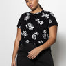 A woman wearing "The Perfect Tee" from the Rose Pop Inclusive Collection by Sanctuary Clothing stands against a plain background. She has her hair pulled back and is wearing black pants. Her right hand rests in her pocket while her left hand hangs by her side.