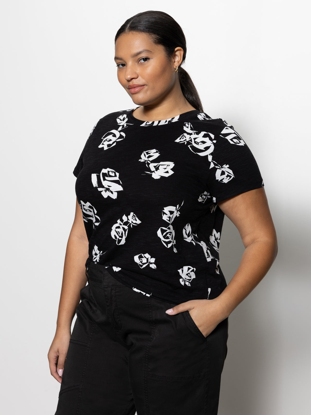 A person with dark hair tied back is wearing the perfect tee from the Rose Pop Inclusive Collection by Sanctuary Clothing. The black short-sleeve top features a striking white rose pattern, which they have paired with black pants. They stand against a plain white background, slightly tilting their head and smiling subtly while keeping one hand in their pocket.