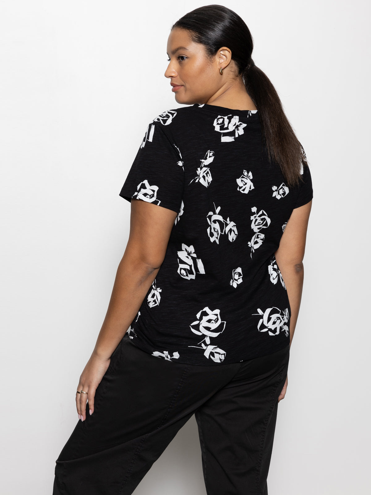 A woman with long hair is standing against a white background, wearing a black t-shirt featuring a white floral pattern from the inclusive collection by Sanctuary Clothing called "the perfect tee rose pop," along with black pants, and she is looking to the left.