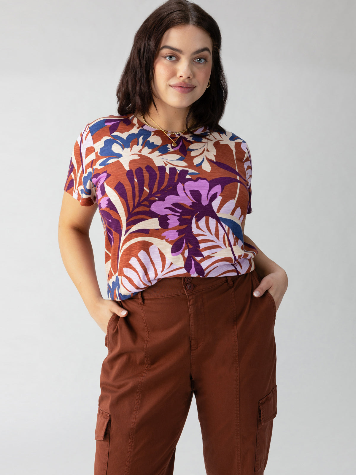 A person with shoulder-length dark hair stands confidently with hands in the pockets of their brown cargo pants. They are wearing Sanctuary Clothing's "The Perfect Tee South Palm Inclusive Collection," a colorful shirt featuring a bold tropical leaf pattern in shades of purple, white, and orange. The background is plain light gray.