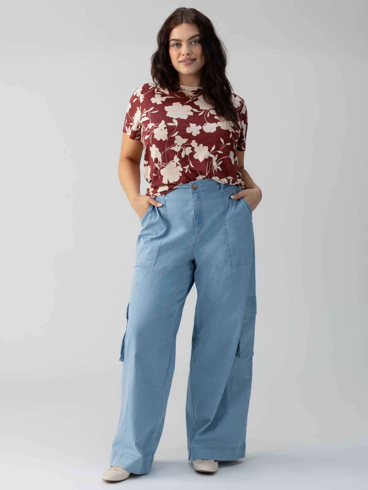 A woman stands against a plain background, wearing The Perfect Tee Warm Vista Inclusive Collection from Sanctuary Clothing, a red floral short-sleeve top. She pairs it with light blue wide-leg pants and has her hands in her pockets while looking at the camera with a relaxed expression. She has long, dark hair and is wearing white shoes.