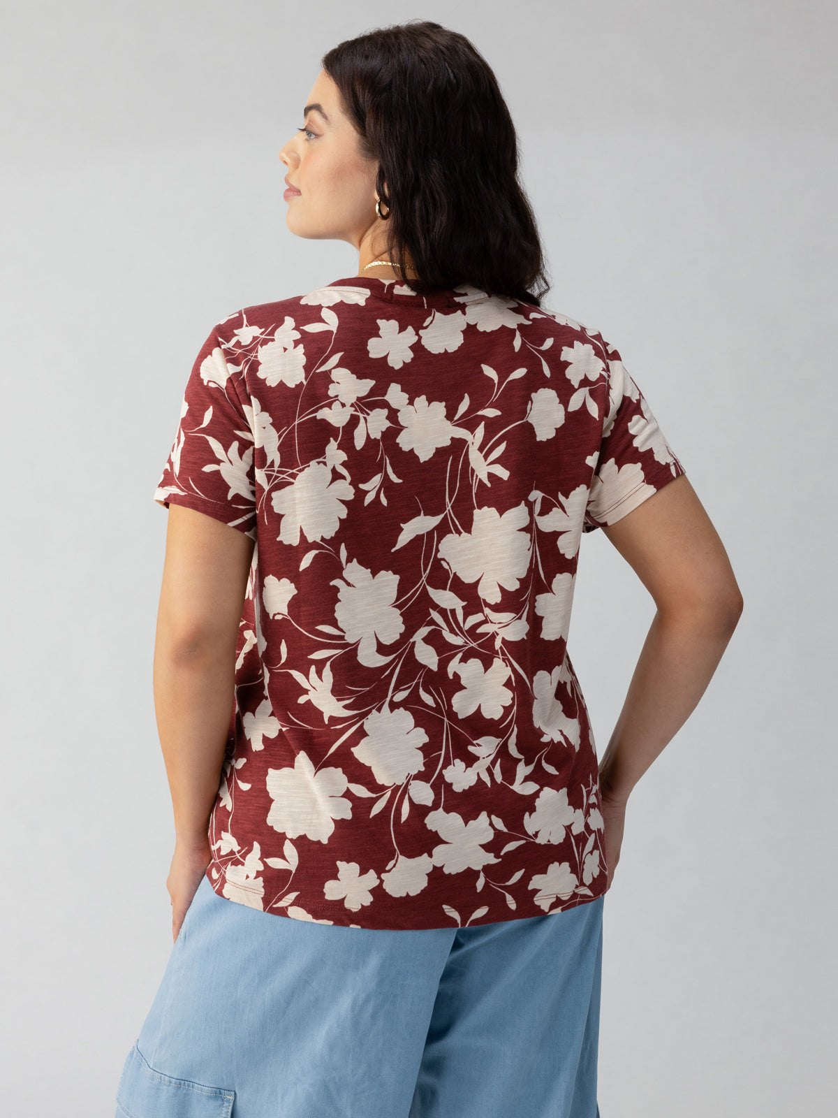 A person with dark hair stands facing away, wearing The Perfect Tee Warm Vista from Sanctuary Clothing's Inclusive Collection, featuring a short-sleeved maroon design with a white floral pattern and paired with light blue pants on a plain gray background. Their right hand is slightly in their pocket while their left arm rests by their side.