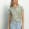 A woman with long blonde hair is wearing "The Perfect Tee Adorned" by Sanctuary Clothing, featuring a green paisley pattern, paired with light blue jeans. She stands against a light background, gazing to her left with a relaxed expression.