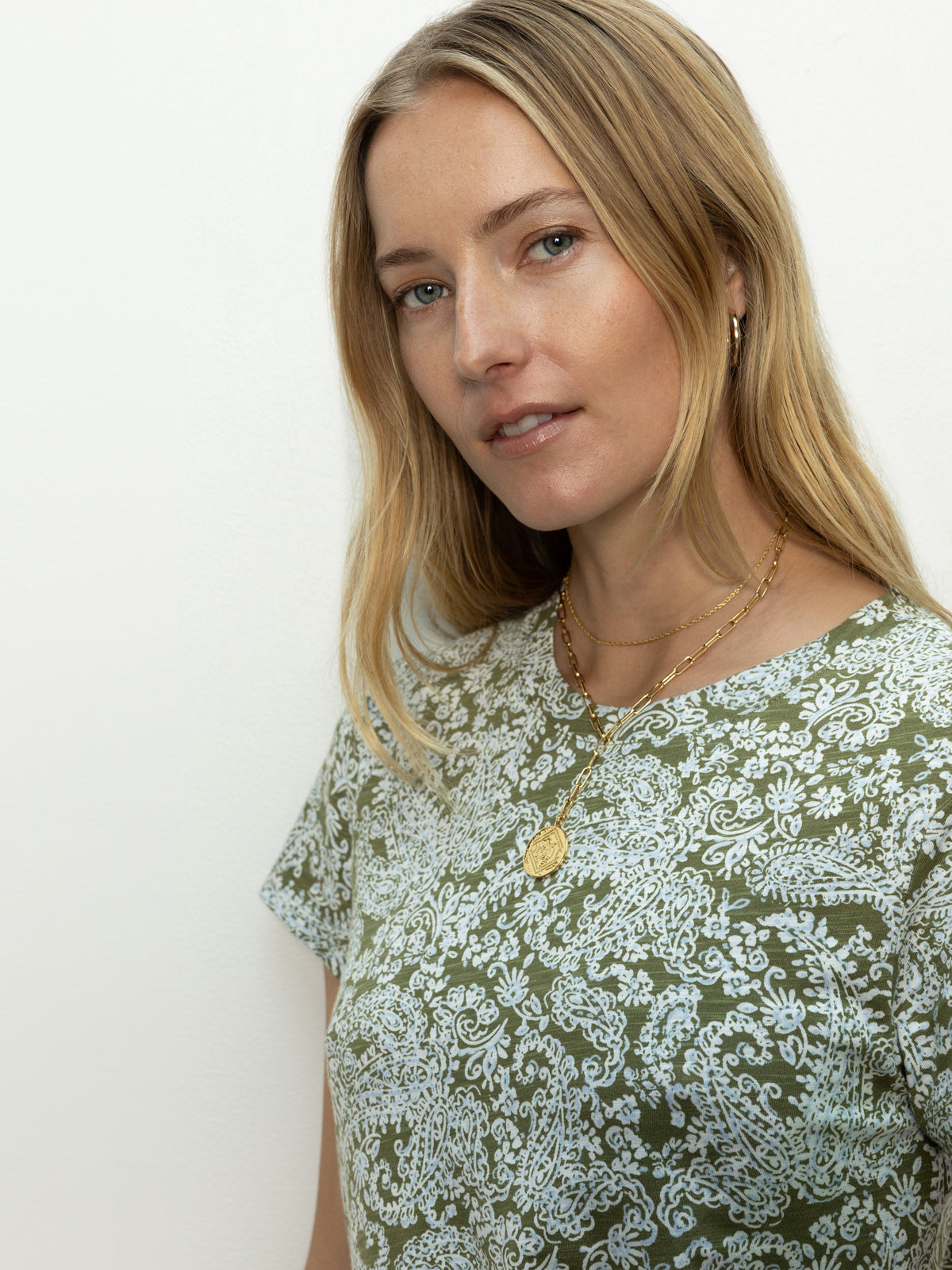 A woman with long blonde hair and gold necklaces wears Sanctuary Clothing's "The Perfect Tee Adorned," a green and white patterned shirt. She has a neutral expression against a plain white background.