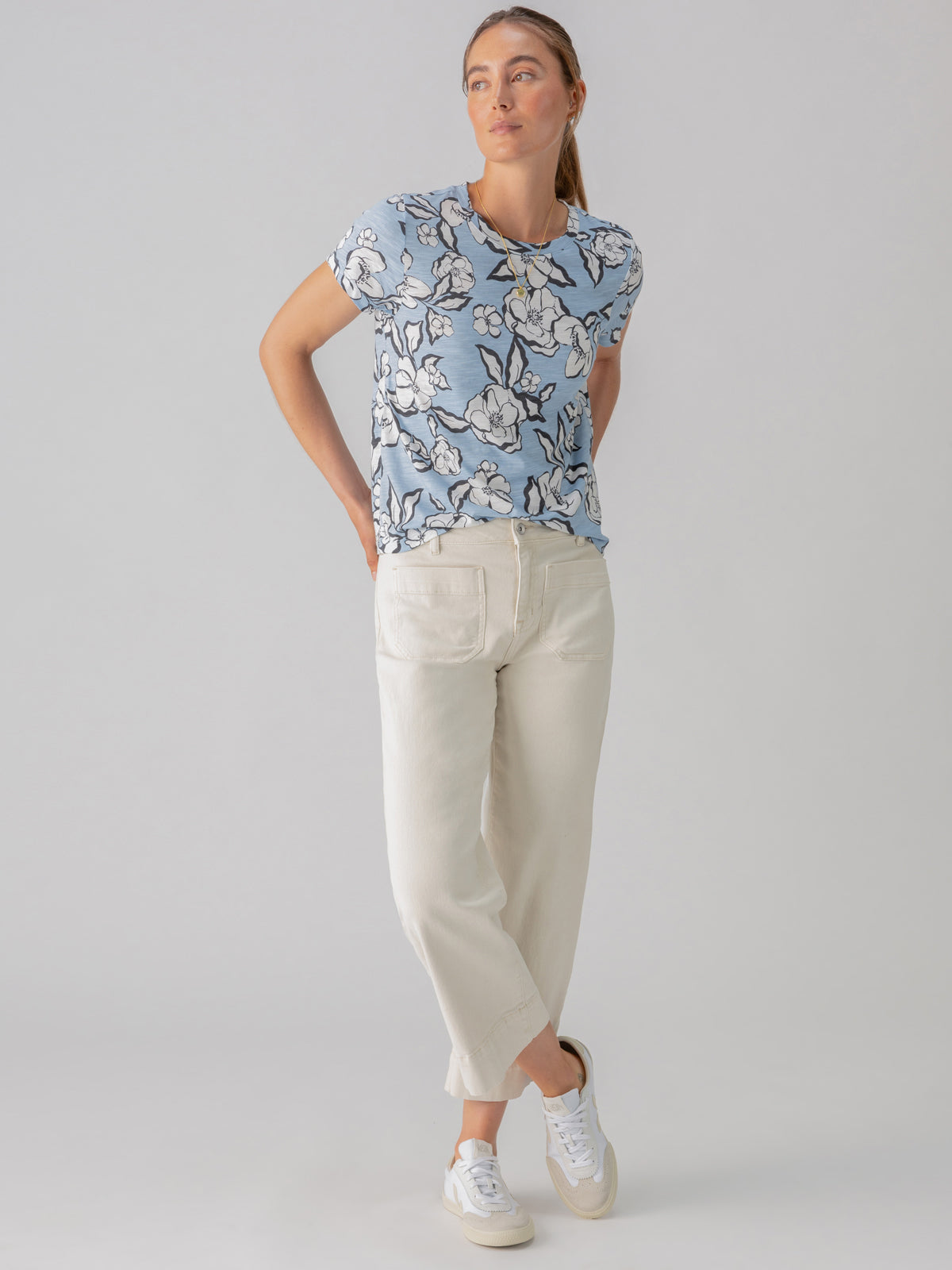 A person stands against a plain background. They are wearing The Perfect Tee Blue Bliss Floral by Sanctuary Clothing, off-white loose-fitting pants, and white sneakers. Their hair is tied back, and they have their hands behind their back while gazing slightly upward to the right.