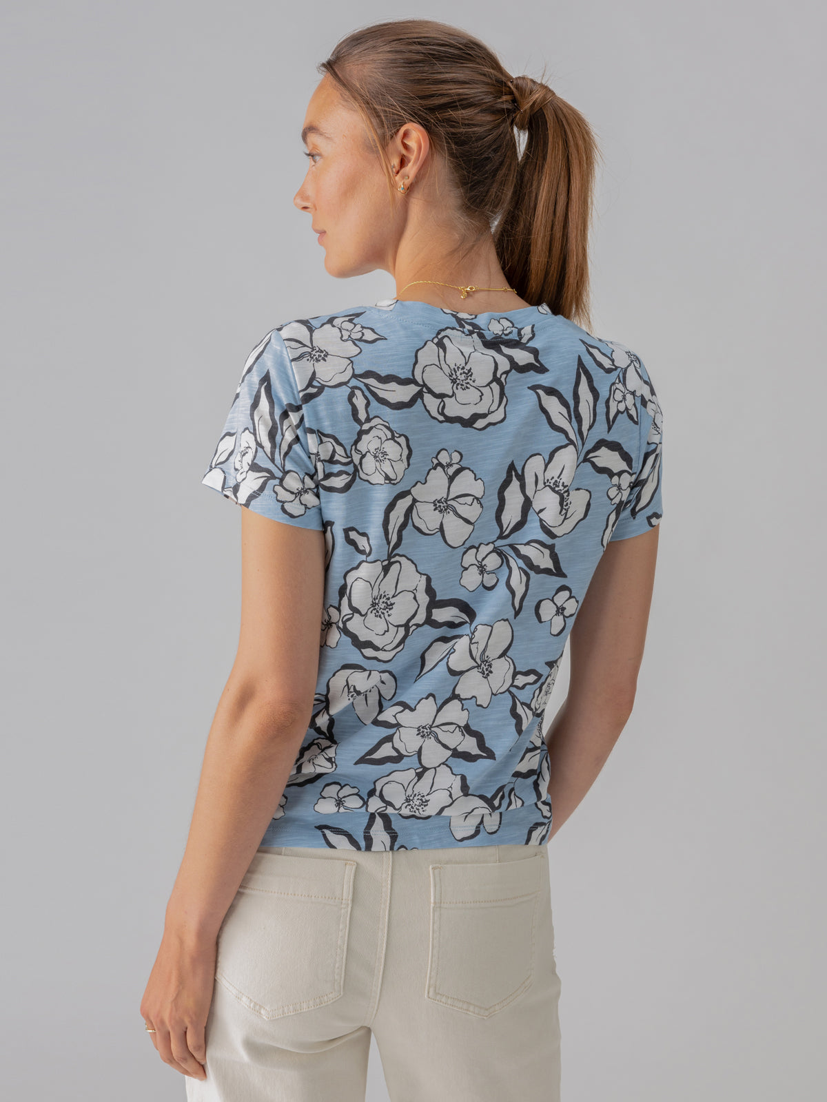 A woman with fair skin and brown hair tied in a ponytail is facing away from the camera, looking to her right. She is wearing Sanctuary Clothing's "The Perfect Tee Blue Bliss Floral" and light-colored pants. The background is a plain, light grey wall.