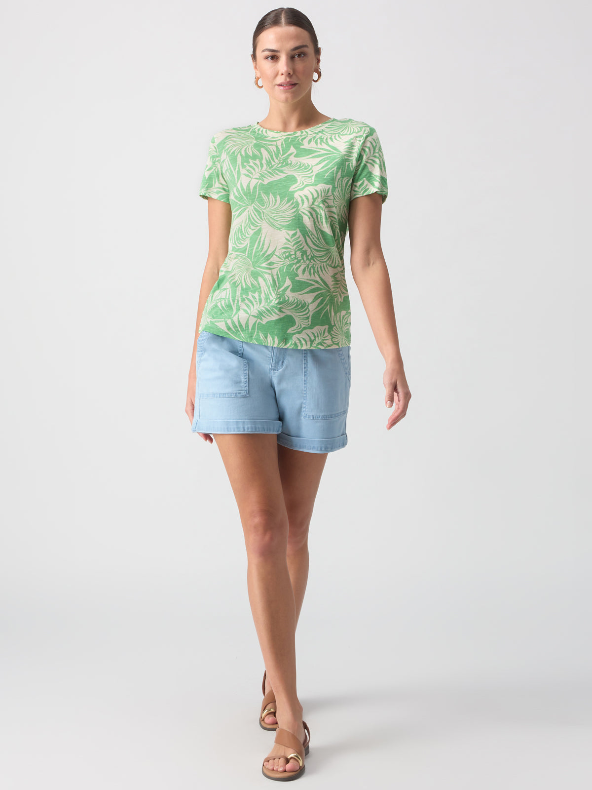 A person with long hair pulled back is standing against a plain background. They are wearing "The Perfect Tee Cool Palm" by Sanctuary Clothing, light blue denim shorts, and sandals. The individual is facing the camera with a neutral expression, hands relaxed at their sides.