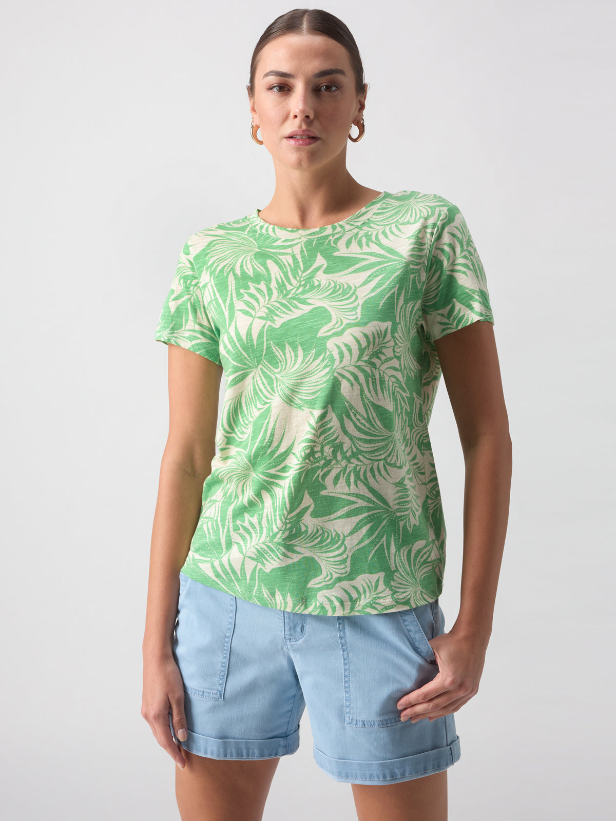 A person with long hair stands confidently against a plain background. They are wearing Sanctuary Clothing's "The Perfect Tee Cool Palm," which features a white leaf pattern, light denim shorts, and hoop earrings. One hand rests in the pocket of their shorts.