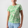 A person with long hair stands confidently against a plain background. They are wearing Sanctuary Clothing's "The Perfect Tee Cool Palm," which features a white leaf pattern, light denim shorts, and hoop earrings. One hand rests in the pocket of their shorts.