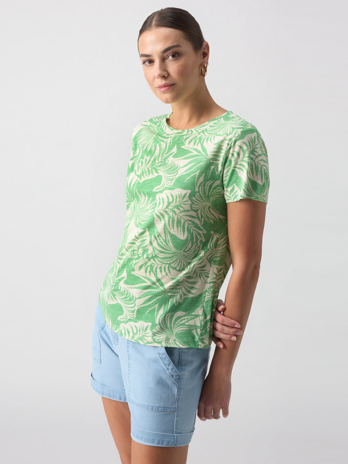 A woman with a slight smile is wearing The Perfect Tee Cool Palm by Sanctuary Clothing, which features a green and white tropical leaf pattern. She pairs it with light blue shorts, has her hair pulled back, and stands against a plain light background.