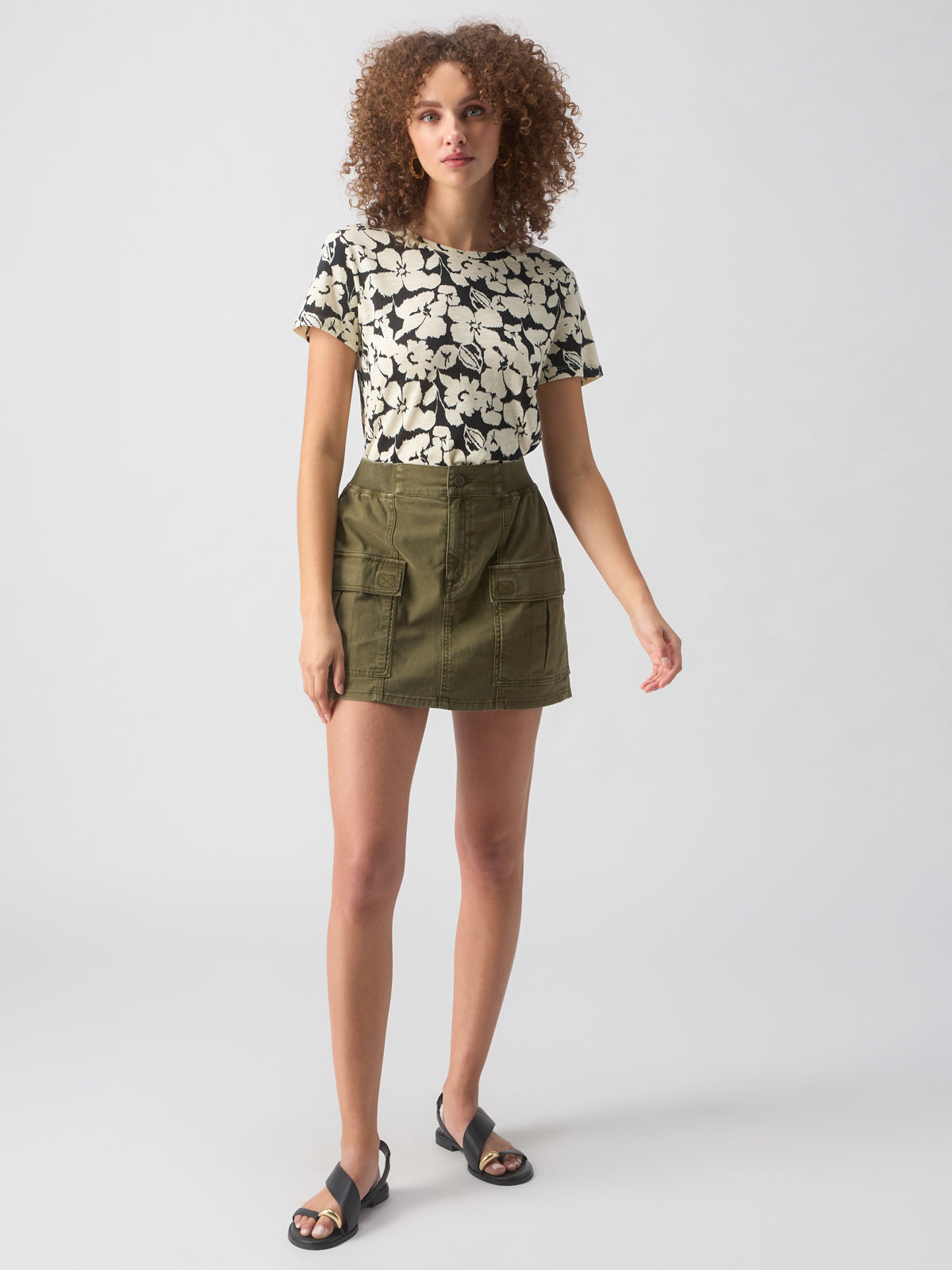 A person with curly hair is wearing Sanctuary Clothing's "The Perfect Tee Echo Blooms," an olive green mini skirt with front pockets, and black sandals, standing against a plain white background.