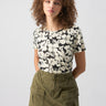 A woman with curly hair wearing The Perfect Tee Echo Blooms by Sanctuary Clothing and an olive green cargo skirt. She has gold hoop earrings and her hands are in her skirt pockets. The background is a plain light grey.