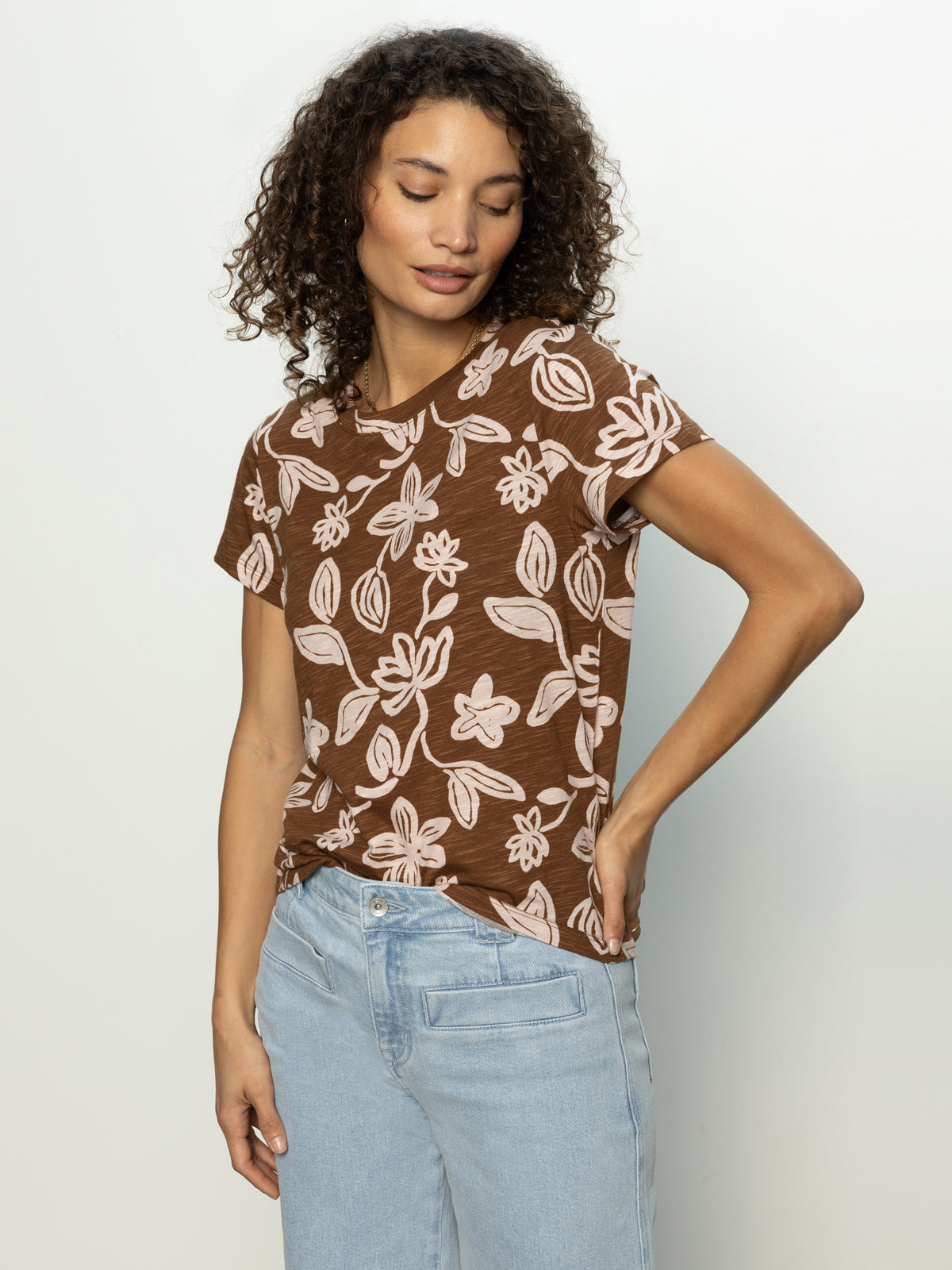 A person with curly hair wearing Sanctuary Clothing's "The Perfect Tee Earth Ivy" and light blue jeans stands against a plain background, displaying a relaxed smile with one hand in their pocket.