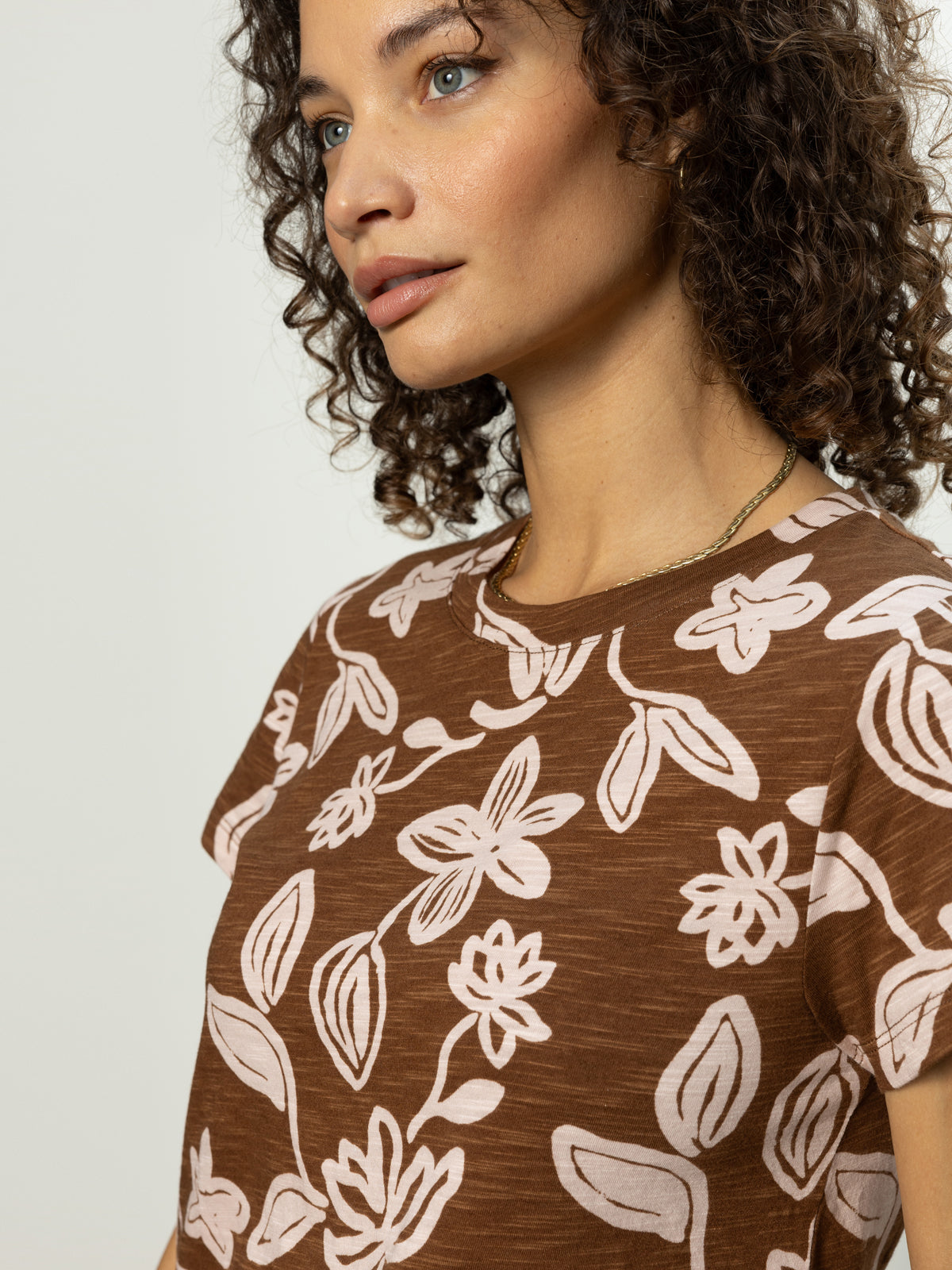 With curly hair, a person wears the perfect tee earth ivy by Sanctuary Clothing, featuring a brown color with a white floral pattern. They glance sideways against a plain background.