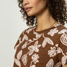 With curly hair, a person wears the perfect tee earth ivy by Sanctuary Clothing, featuring a brown color with a white floral pattern. They glance sideways against a plain background.