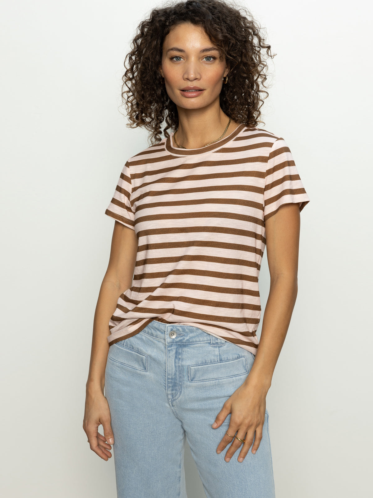 A woman in Sanctuary Clothing's "the perfect tee earth pink sepia stripe" and light blue jeans stands against a plain white backdrop. With curly hair, she gazes neutrally at the camera.