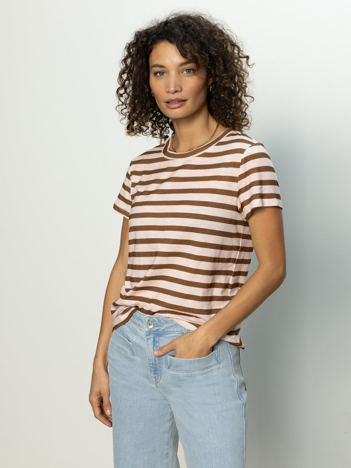 A person with curly hair is wearing the Sanctuary Clothing's "the perfect tee earth pink sepia stripe" paired with light blue jeans. They stand against a plain background, gazing at the camera with a neutral expression.