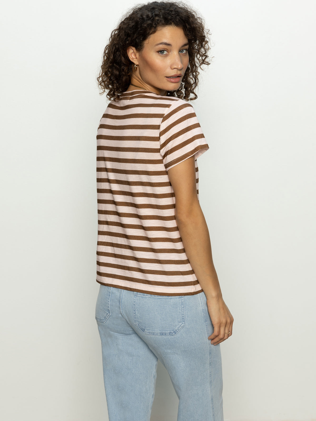 A woman with curly hair models Sanctuary Clothing's "the perfect tee earth pink sepia stripe," paired with light blue jeans, as she looks over her shoulder against a plain background.