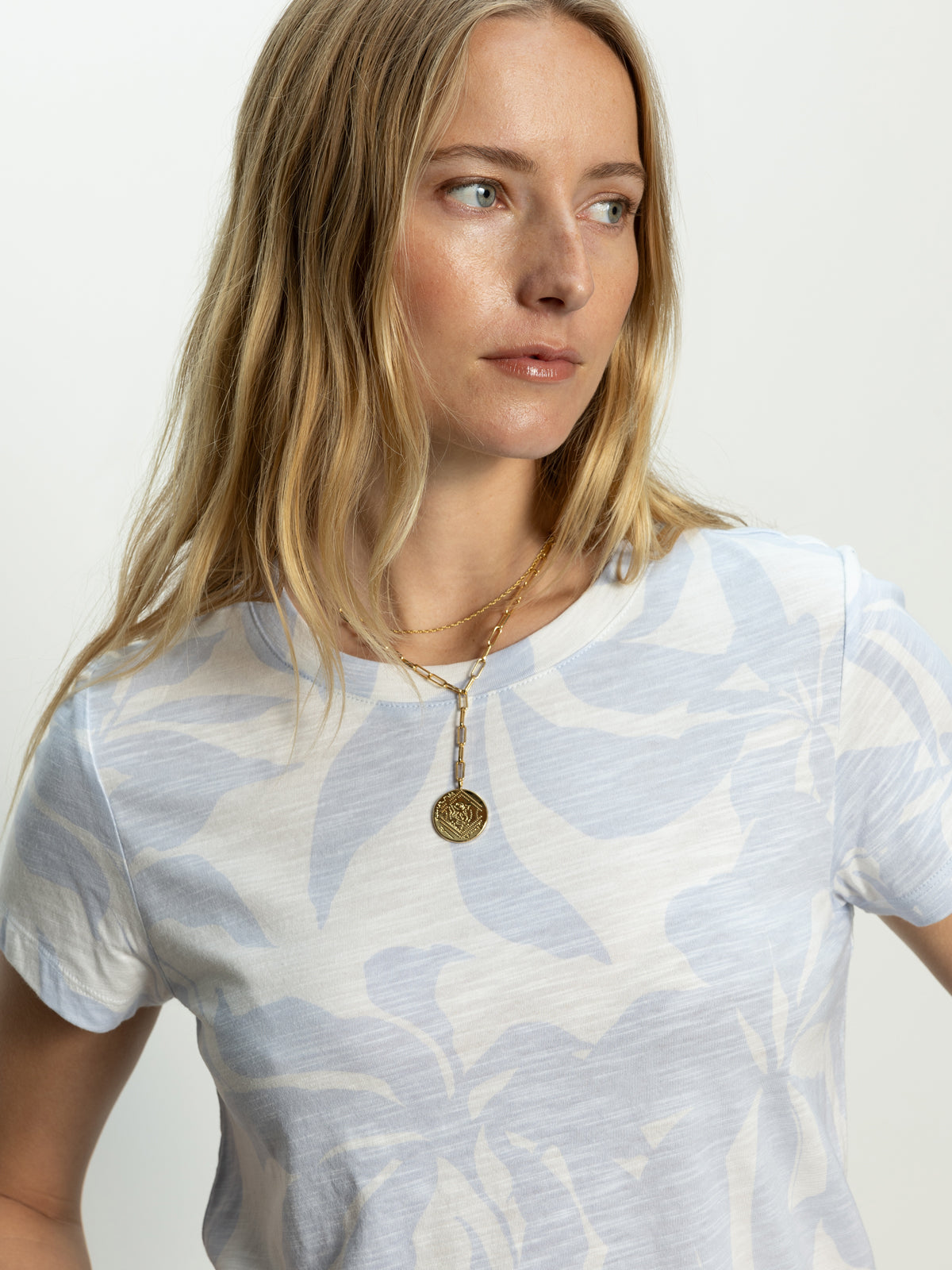 A woman with long blonde hair wears Sanctuary Clothing's "the perfect tee floral bliss," a white and light blue patterned shirt. She gazes to the side and sports a layered gold necklace with a round pendant.