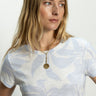 A woman with long blonde hair wears Sanctuary Clothing's "the perfect tee floral bliss," a white and light blue patterned shirt. She gazes to the side and sports a layered gold necklace with a round pendant.